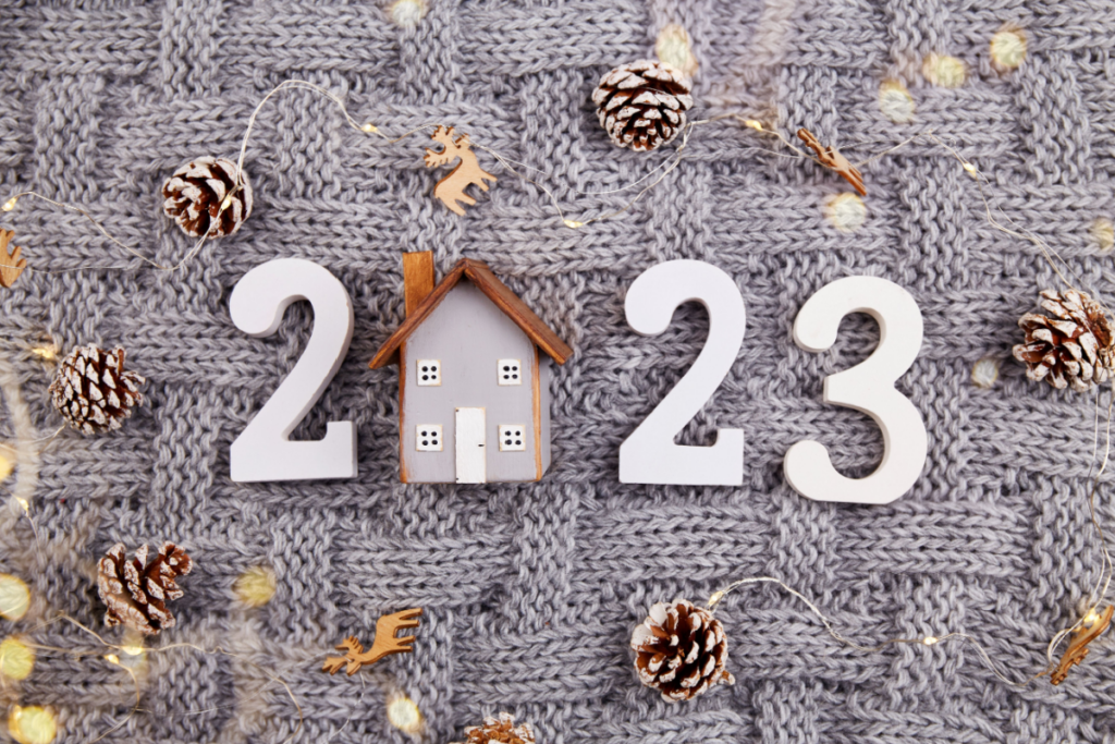 Tecido de lá com número 2023 e casinha de madeira no lugar do 0. Pinhas, renas de madeiras e luzes de natal em volta