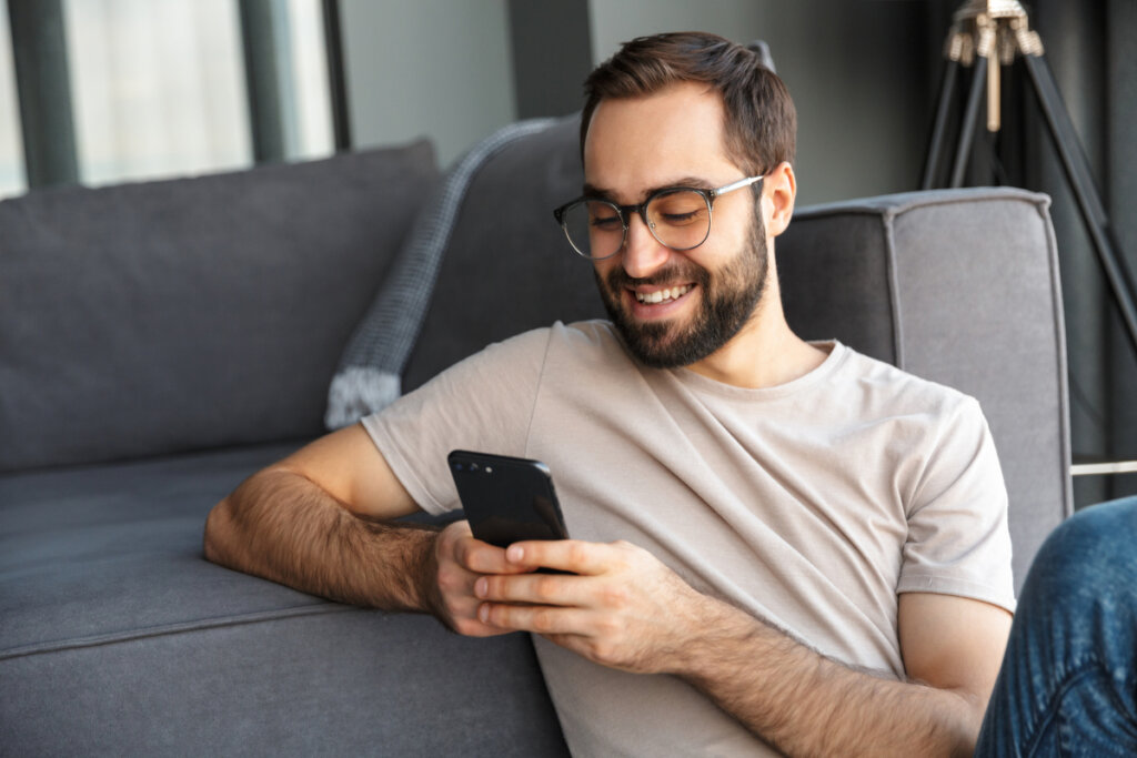 Homem de óculos sentado sorrindo e mexendo no celular 