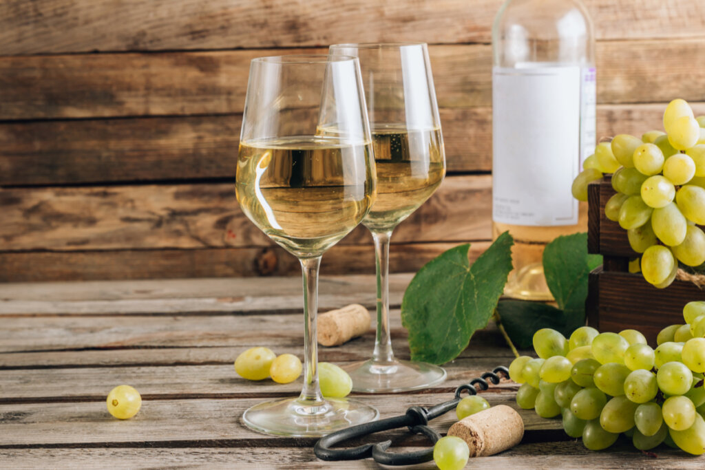 Duas taças de vinho branco, uvas verdes, saca-rolhas e garrafa de vinho ao fundo