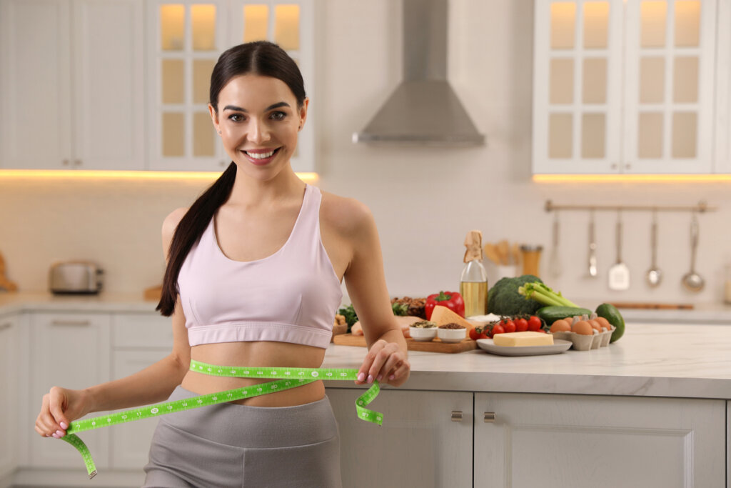 Mulher medindo a cintura com fita métrica e alimentos saudáveis em cima da mesa ao fundo