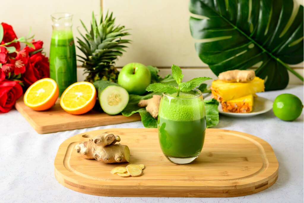 Copo de suco verde com gengibre ao lado em cima da tábua de madeira e frutas ao fundo