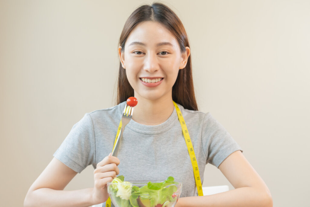 Mulher sentada sorrindo segurando um garfo com tomate e um recipiente com salada