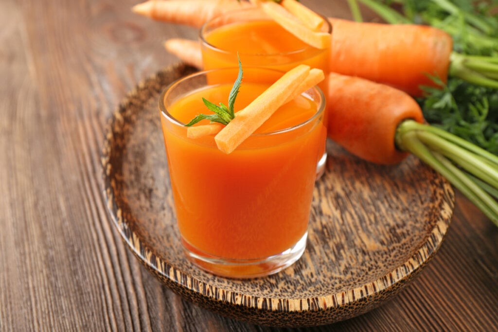 Dois copos com suco de cenoura e cenouras inteiras ao fundo em cima da mesa de madeira