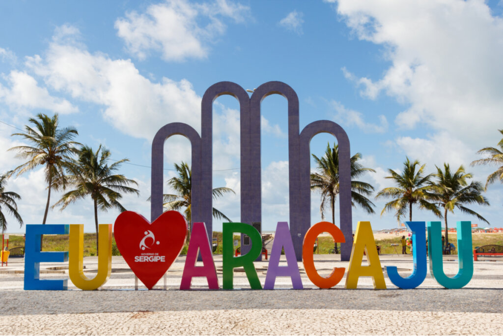 Letreiro com "eu amo Aracaju", céu azul e coqueiros ao fundo