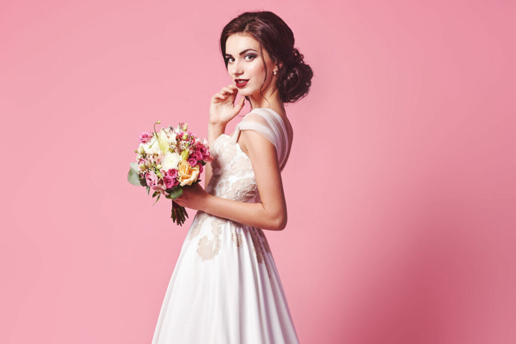 Mulher com vestido de noiva segurando buquê de rosas em frente a um fundo rosa