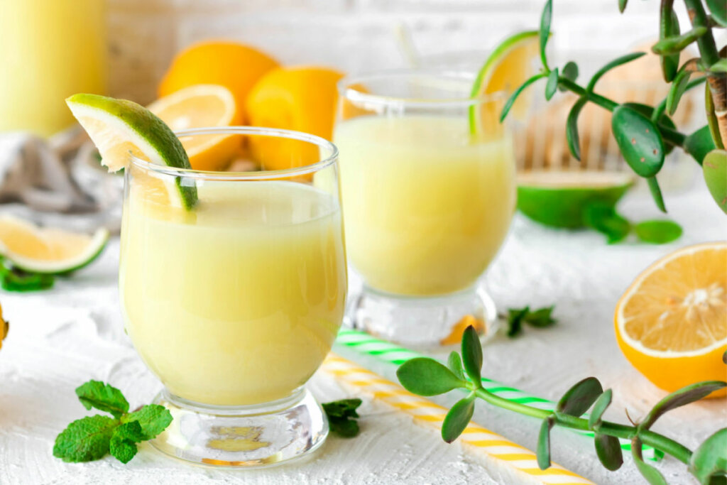 Suco amarelo em copa de vidro com rodela de laranja na borda e frutas, plantas e canudos em volta