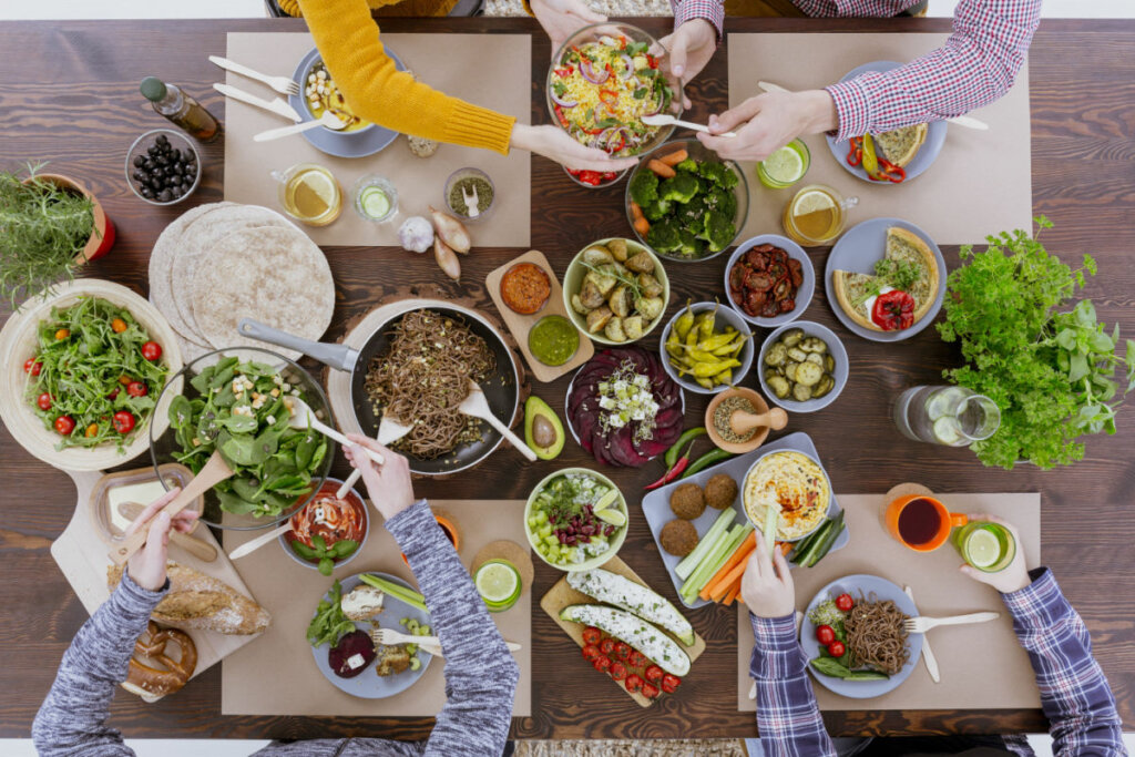 Vista de cima de uma mesa repleta de comidas e vegetais com quatro pessoas se servindo