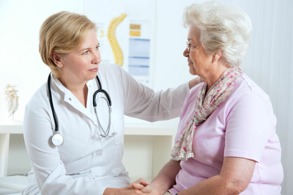 Médica sentada de frente para idosa com a mão no ombro dela