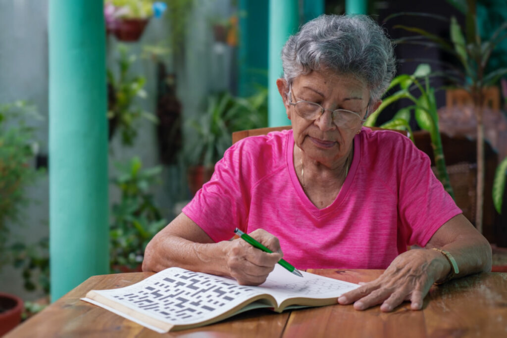 Mulher idosa sentada em mesa fazendo palavra cruzada em livro