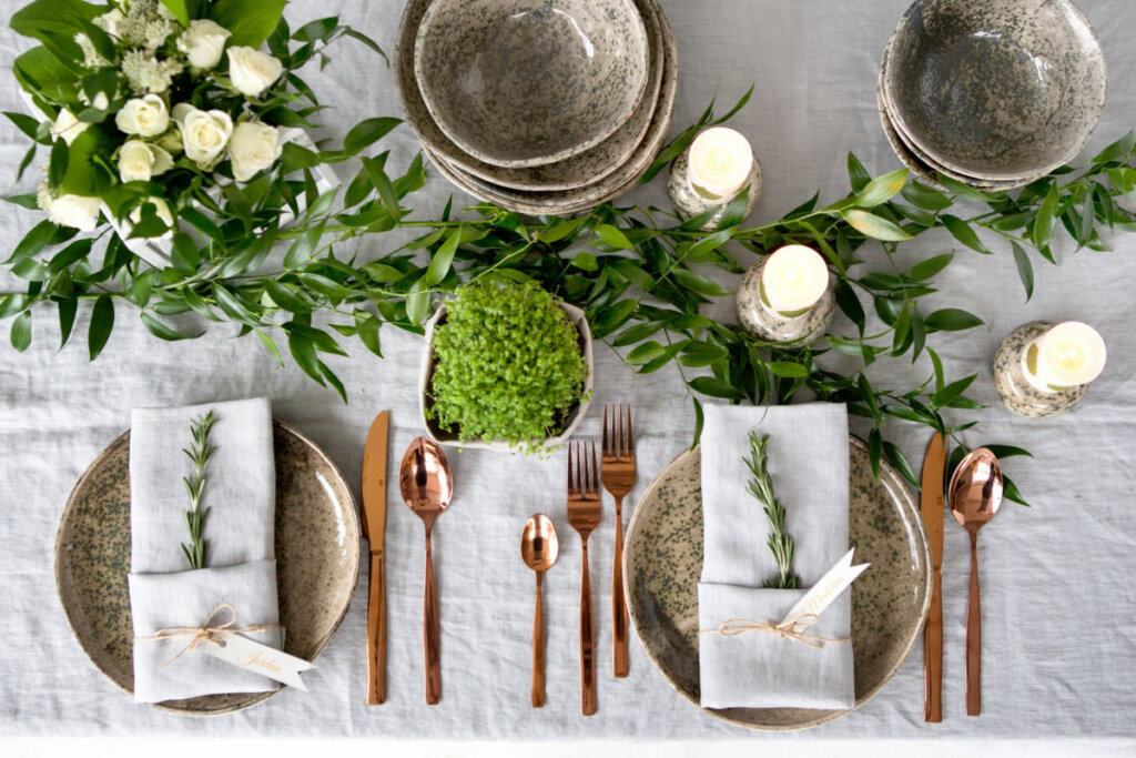 Mesa com pratos de cerâmica, guardanapos brancos e talheres de metal. Mesa tem plantas e velas 