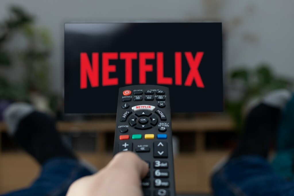 Uma mão segurando controle e logo da Netflix em televisão