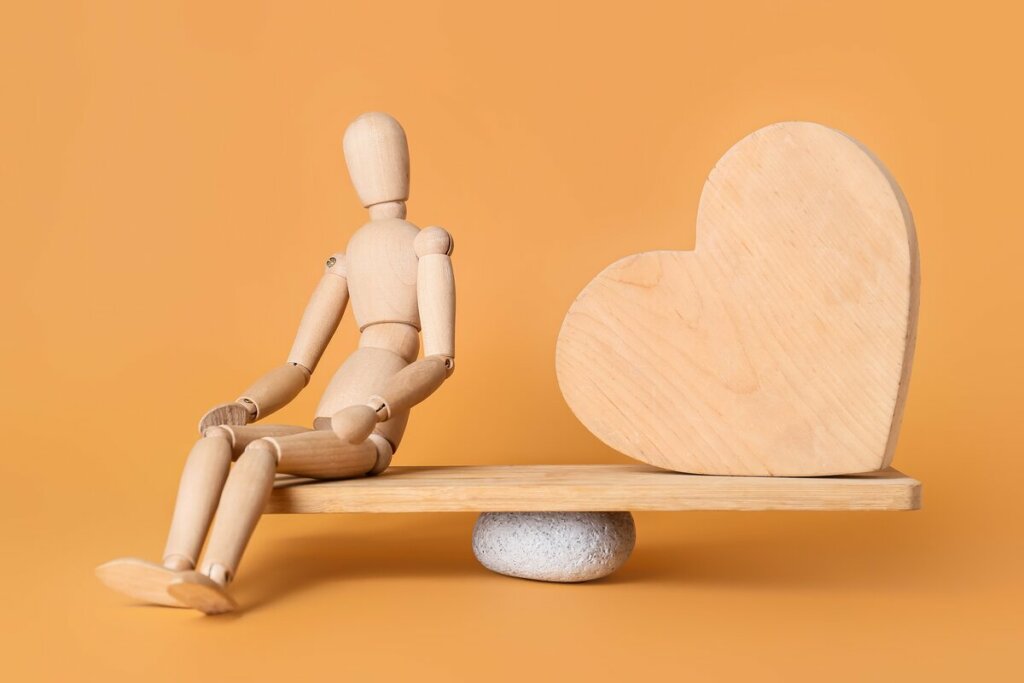 Boneco de madeira equilibrando uma balança com um coração de madeira em frente a um fundo amarelo