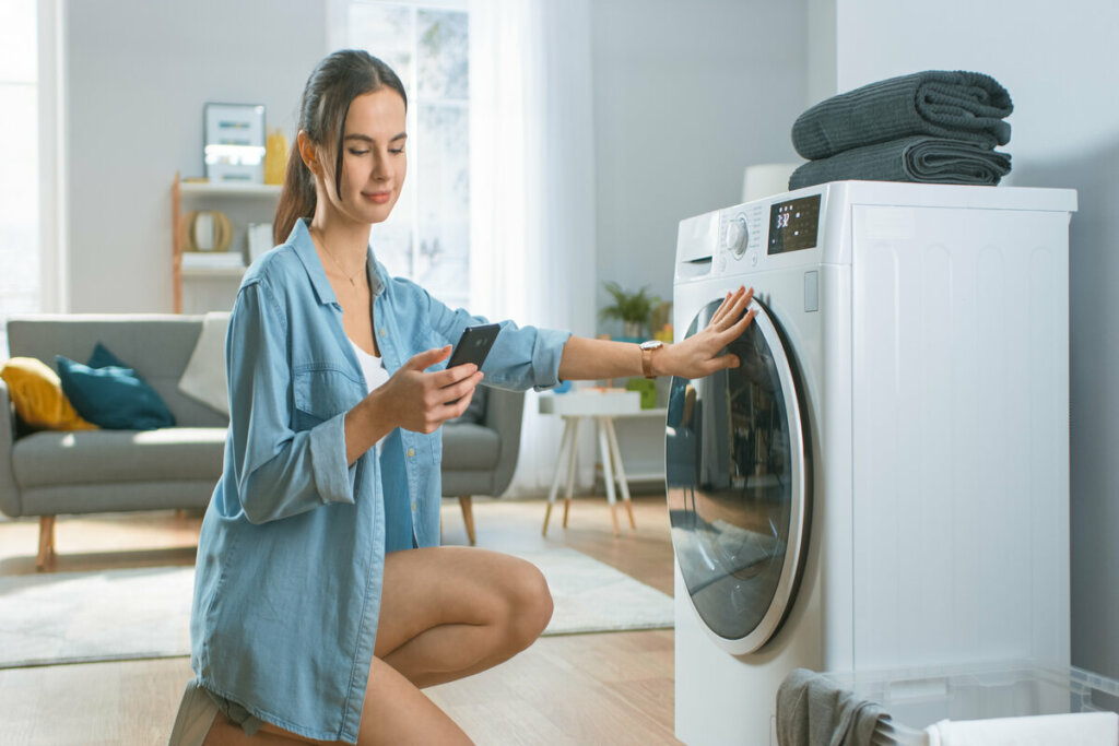 Mulher olhando o celular com a mão em frente a uma máquina de lavar roupa