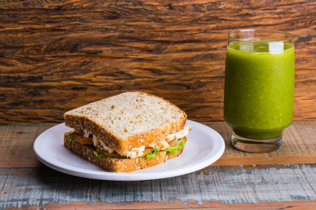 Sanduíche com frango, alface e tomate em cima de um prato branco com copo de suco verde ao lado