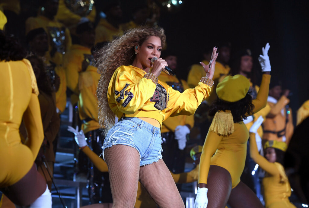 Artista Beyoncé de moletom amarelo e shorts jeans cantando em palco.