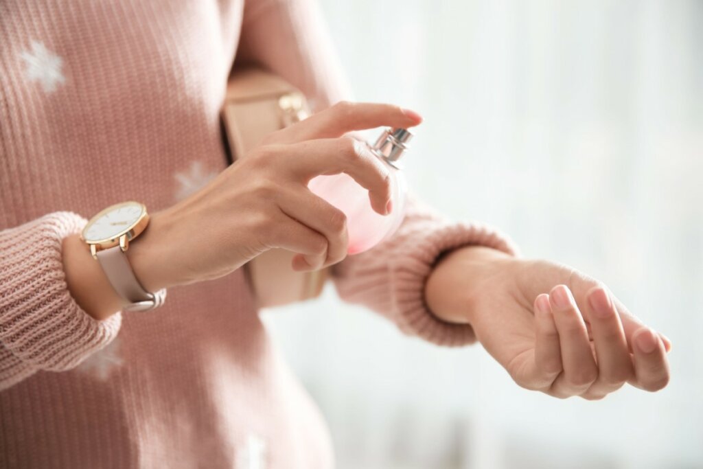 Na imagem, uma mulher aplica perfume no pulso esquerdo. Ela está com uma blusa de lã rosa e um relógio no pulso direito.