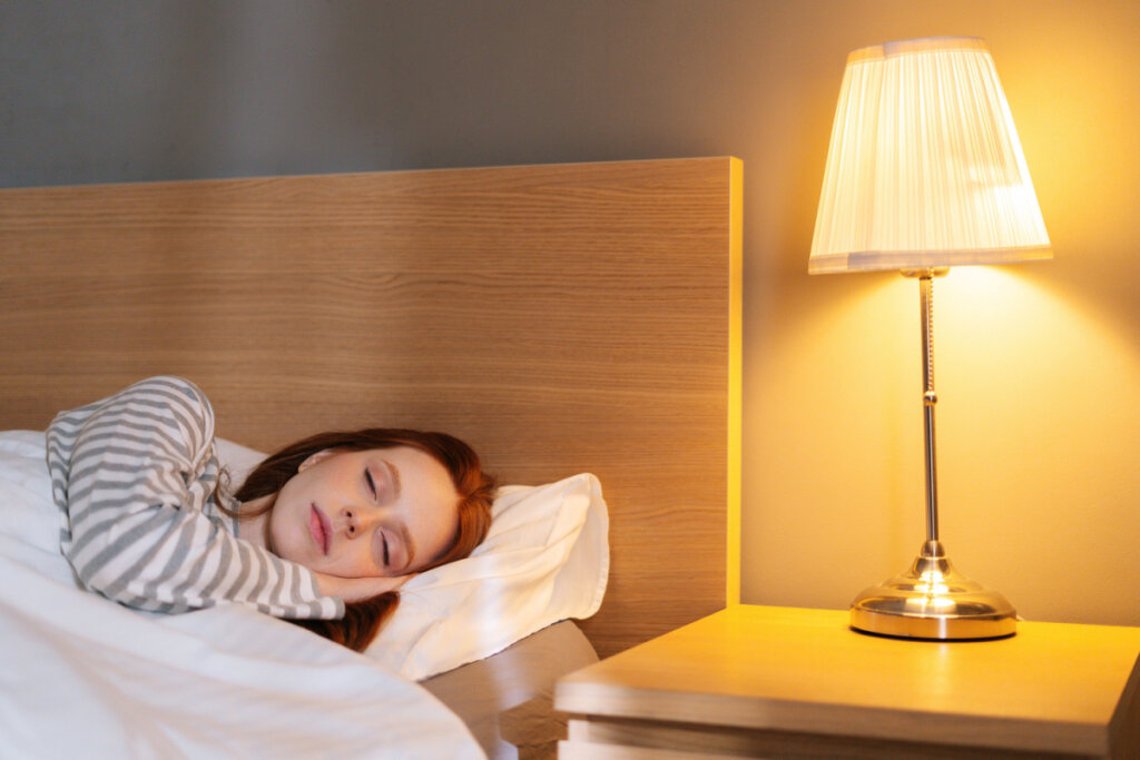 Na imagem, mulher dorme em uma cama com cabeceira de madeira. Ao lado, uma mesa de canto e um abajur com lâmpada de tom quente e amarela