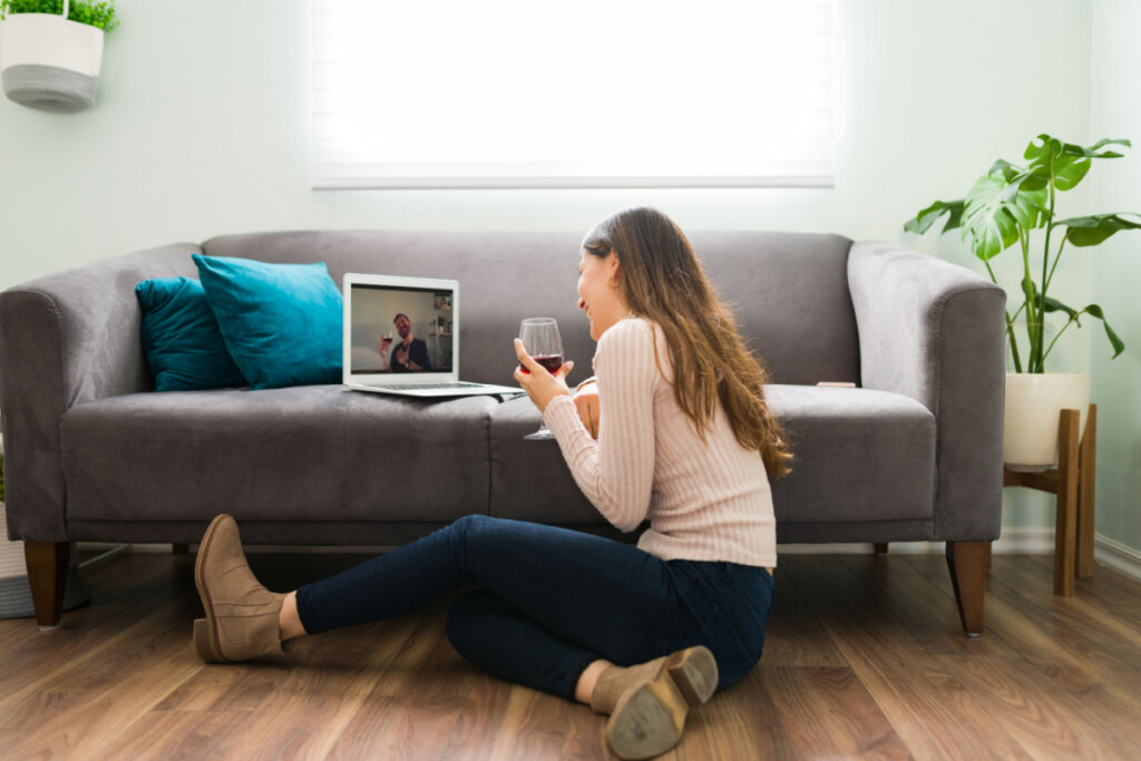 Mulher sentada no chão da sala com uma taça de vinho conversando com o parceiro por vídeo em um computador, do outro lado da tela; ele também bebe vinho. Ambos sorriem. 
