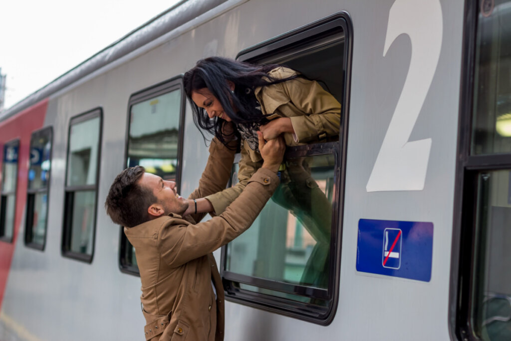 Na imagem, casal se despede em estação de trem. Os dois usam casaco bege e dão as mãos pela janela