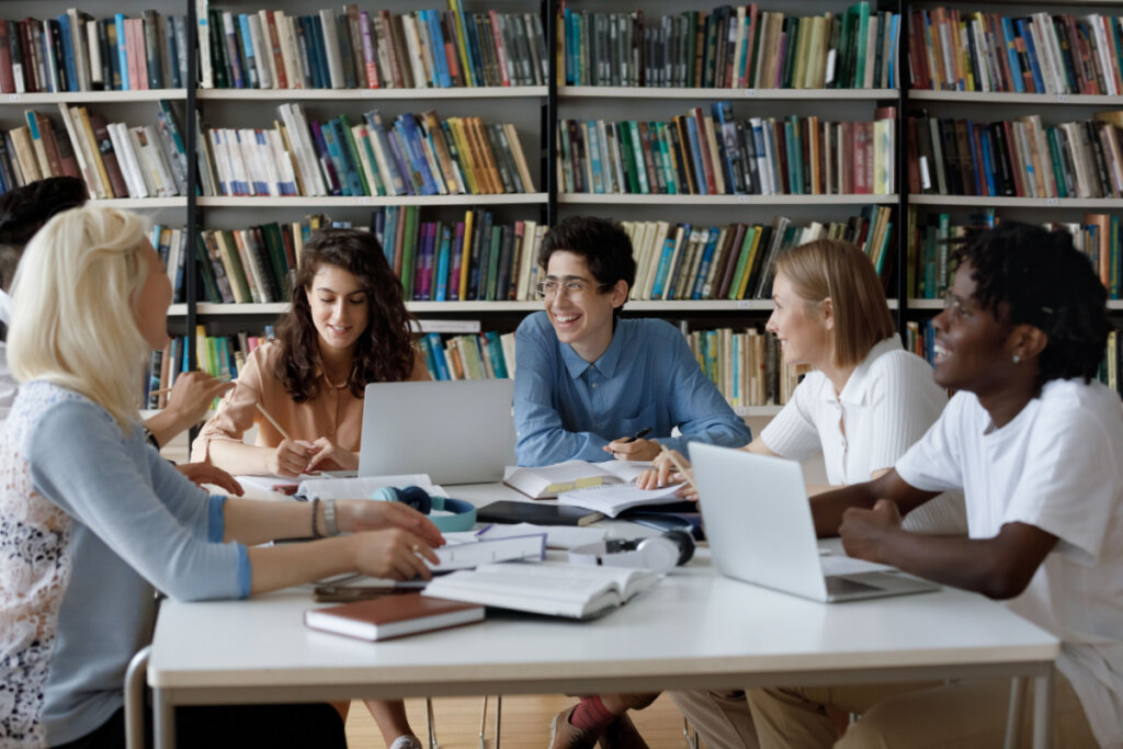 Na imagem, um grupo de seis pessoas está sentado em uma mesa de estudos com livros, computador, fone de ouvido e cadernos espalhados. As pessoas sorriem. 