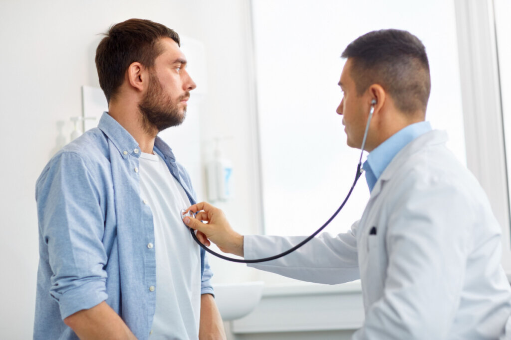 Na foto, um médico examina outro homem, de barba, com um estetoscópio para conferir os batimentos cardíacos. Ambos estão com a expressão facial séria