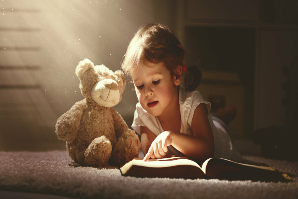 Menina deitada lendo livro para um urso de pelúcia