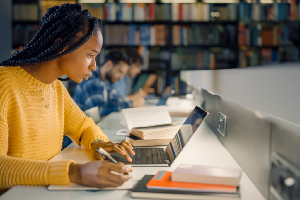 Na imagem, jovem estudante em uma biblioteca escrevendo no computador e em um caderno concentrada