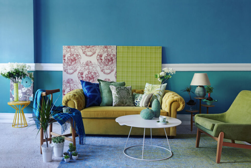 Sala de estar com sofá amarelo, almofadas coloridas, poltrona verde, parede azul e verde e outros elementos coloridos