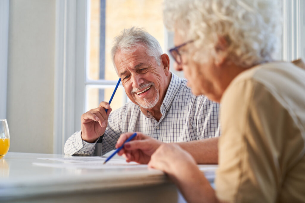 Homem e mulher idosos estão sentados em uma mesa com papéis e canetas realizando exercícios para estimular a cognição e memória