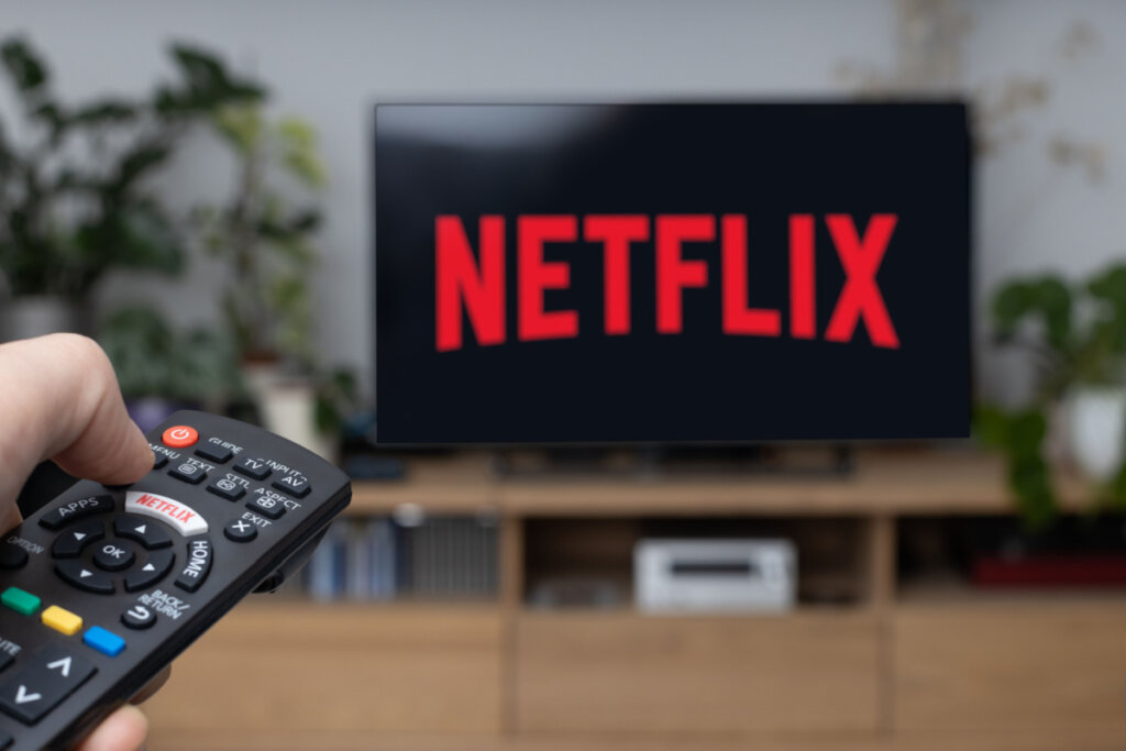 TV com logo da Netflix em rack marrom. Mão com controle remoto apontado para a TV