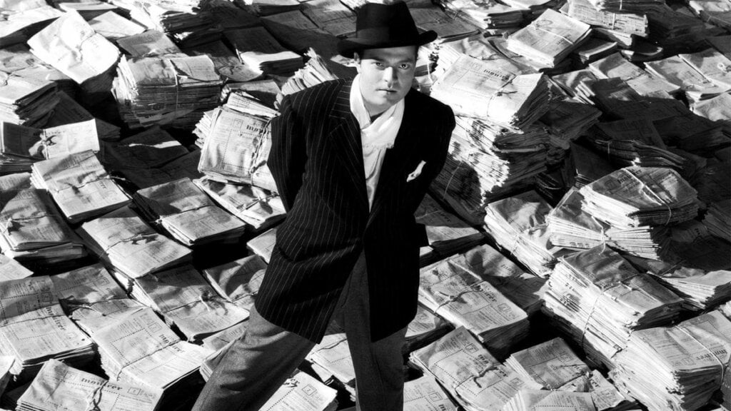 Imagem do filme Cidadão Kane. Um homem em volta de diversas pilhas de jornais impressos
