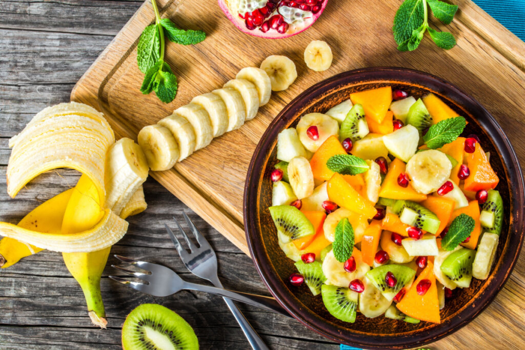 Bowl com variedade de frutas, como manga e kiwi. Em tábua de madeira, banana cortada em fatias.