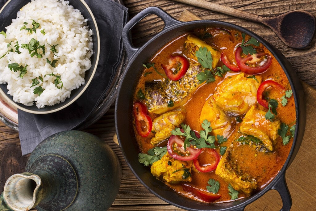 Mesa com uma travessa de cerâmica arroz branco, uma panela grande de ferro com moqueca de peixe e um jarro de cerâmica cinza