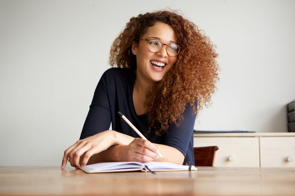 Mulher de óculos sorrindo e segurando um lápis em cima de um caderno 