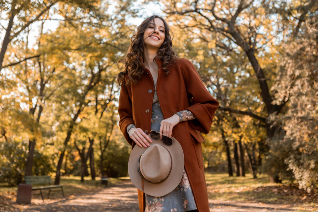 Mulher elegante segurando chapéu e óculos de sol andando no parque em meio a folhas de outono. Ela usa um casaco quente de outono e acessórios.