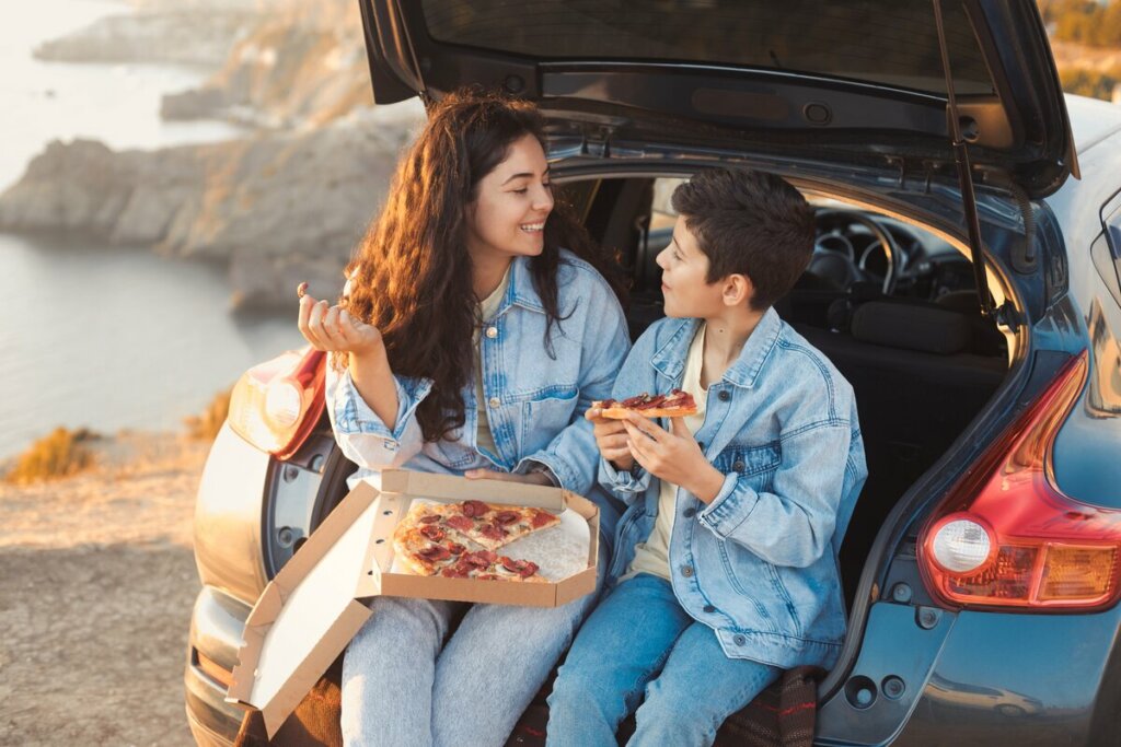 Mulher e menino sentados no porta malas de um carro comendo pizza