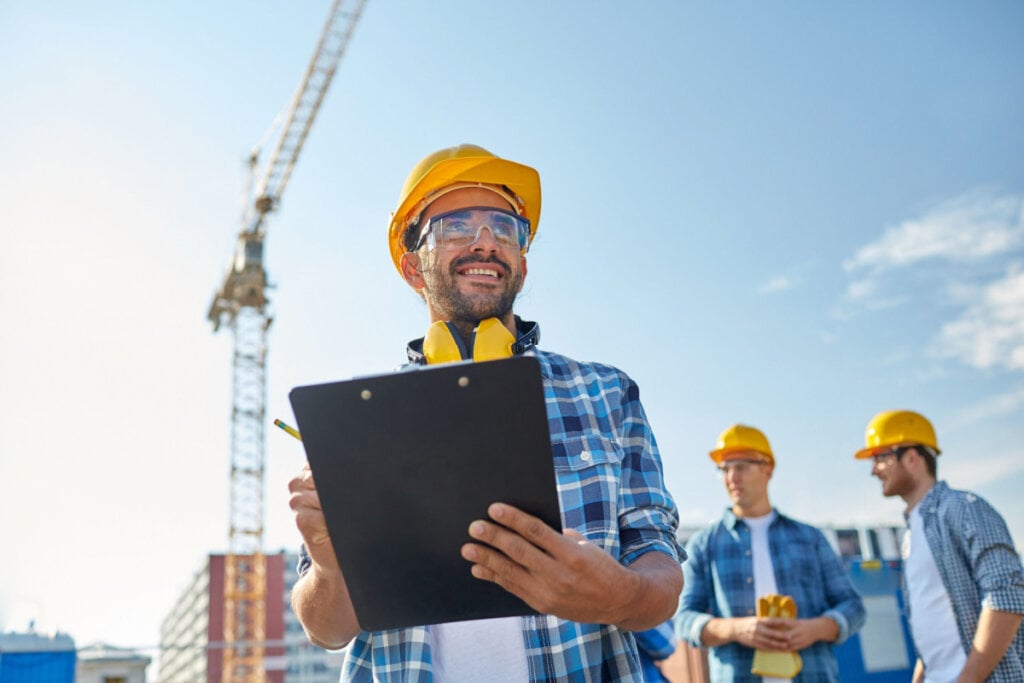 Chefe de construção com prancheta na mão; logo atrás tem mais outros dois trabalhadores em pé conversando; na imagem, todos estão de blusa xadrez azul e com capacete amarelo