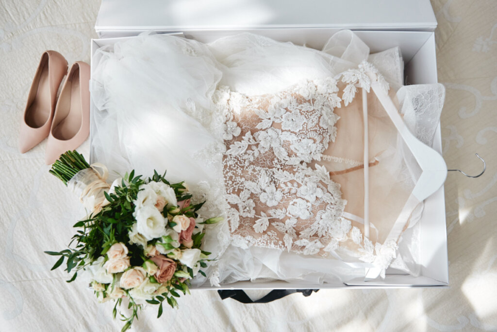 Vestido de noiva em cabide dobrado, com buquê em cima e sapatos de salto ao lado.