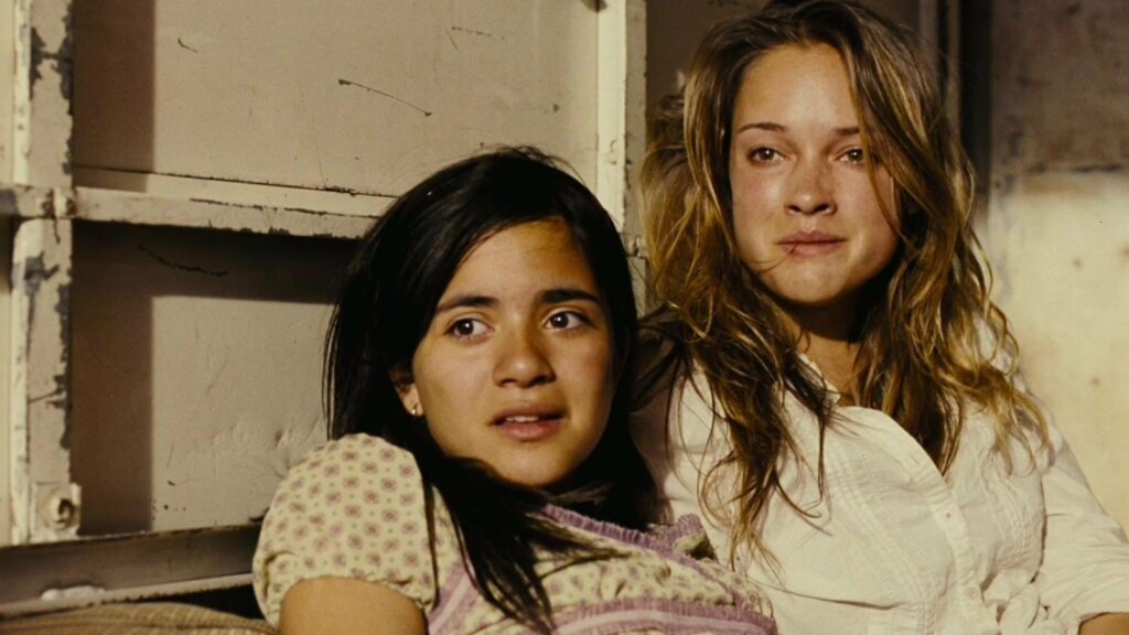 Imagem do filme "Tráfico, Bem-vindo à América". Duas meninas assustadas e com marcas de machucado, encostadas em parede branca descascada.