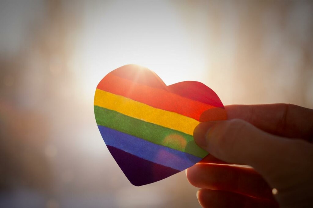 Uma mão segurando um coração colorido com as cores da bandeira LGBTQIA+