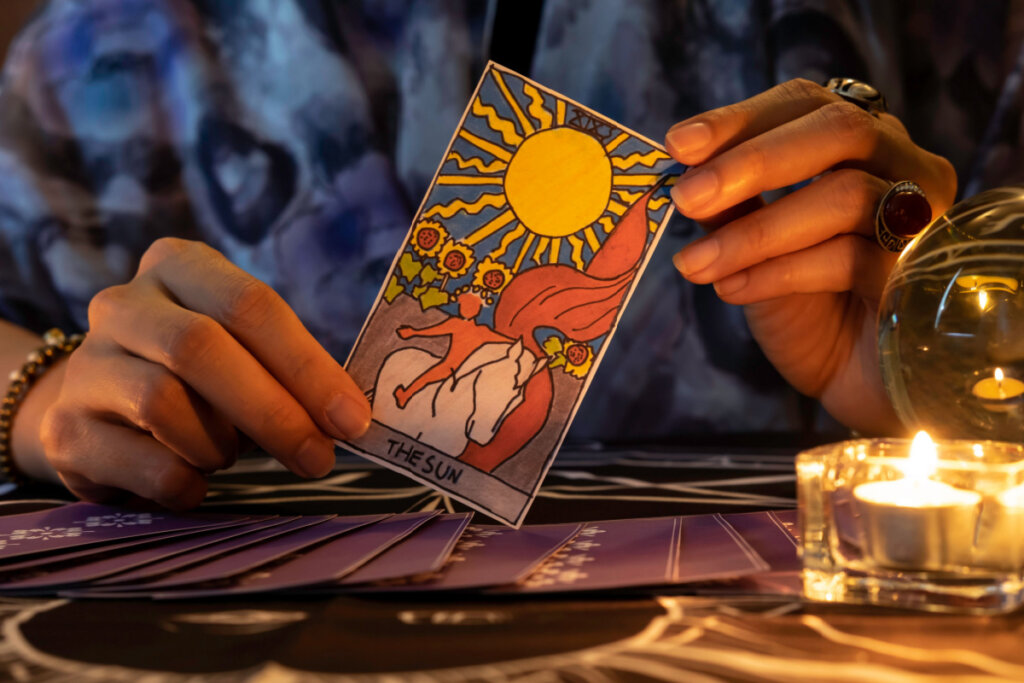 Cartomante de mãos segurando o cartão THE SUN e cartas de tarô na mesa perto de velas acesas à luz de velas