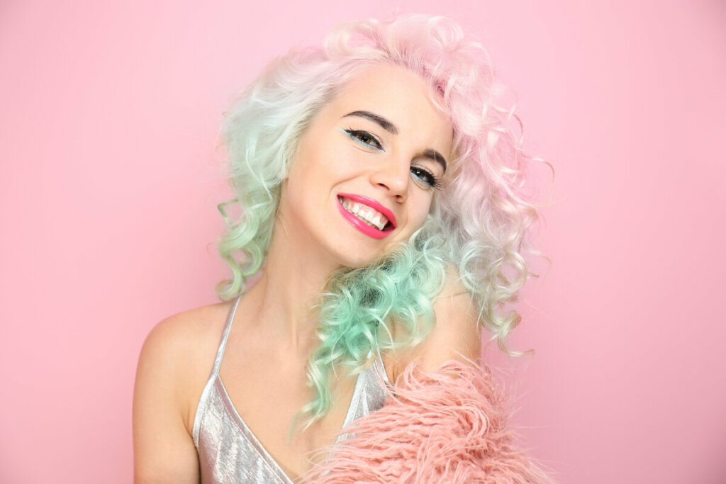 Mulher com cabelo rosa e verde sorrindo e posando para foto em frente a um fundo rosa