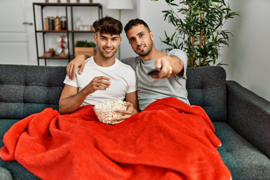 Dois homens abraçados assistindo filme com cobertor e pipoca.