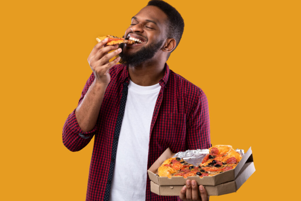 Homem comendo fatia de pizza enquanto segura caixa com o restante do alimento.