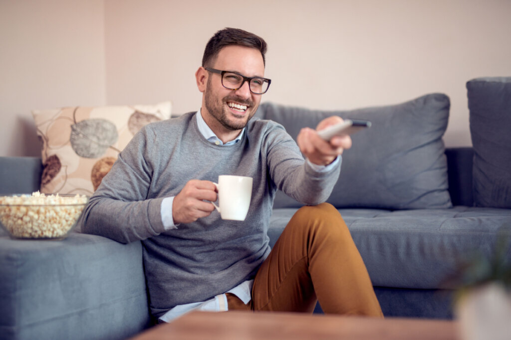 Homem feliz comendo pipoca enquanto está sentado em um sofá em casa e assistindo à TV.