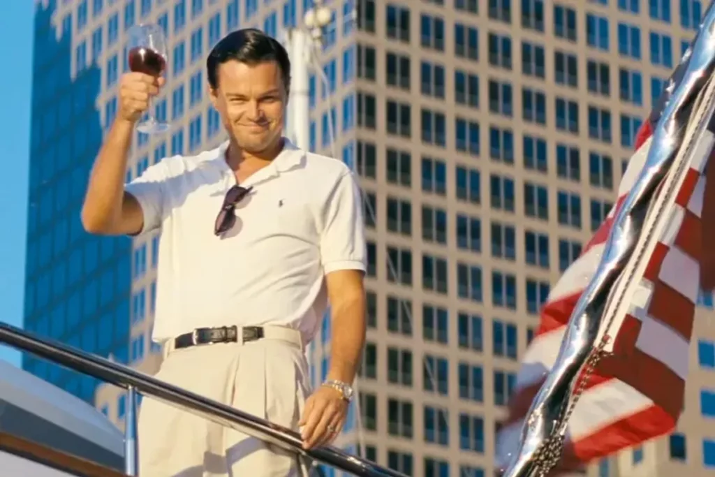 Cena do filme O Lobo de Wall Street, com Leonardo DiCaprio segurando taça de vinho, sorrindo e de roupa branca ao lado de bandeira dos Estados Unidos.