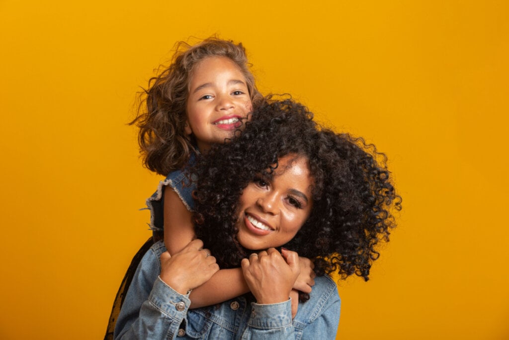 Mãe com filha apoiada nas costas em fundo amarelo