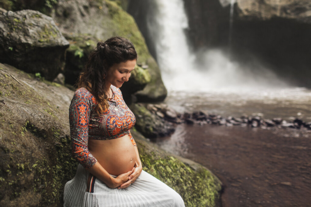Mulher grávida em cachoeira com as mãos na barriga e olhando para baixo, sorrindo.
