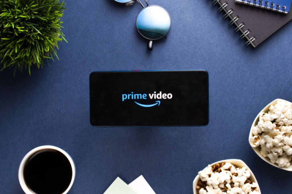Logo do Prime Video em celular rodeado com pipoca, café, planta, óculos de sol e outros adereços em fundo azul..