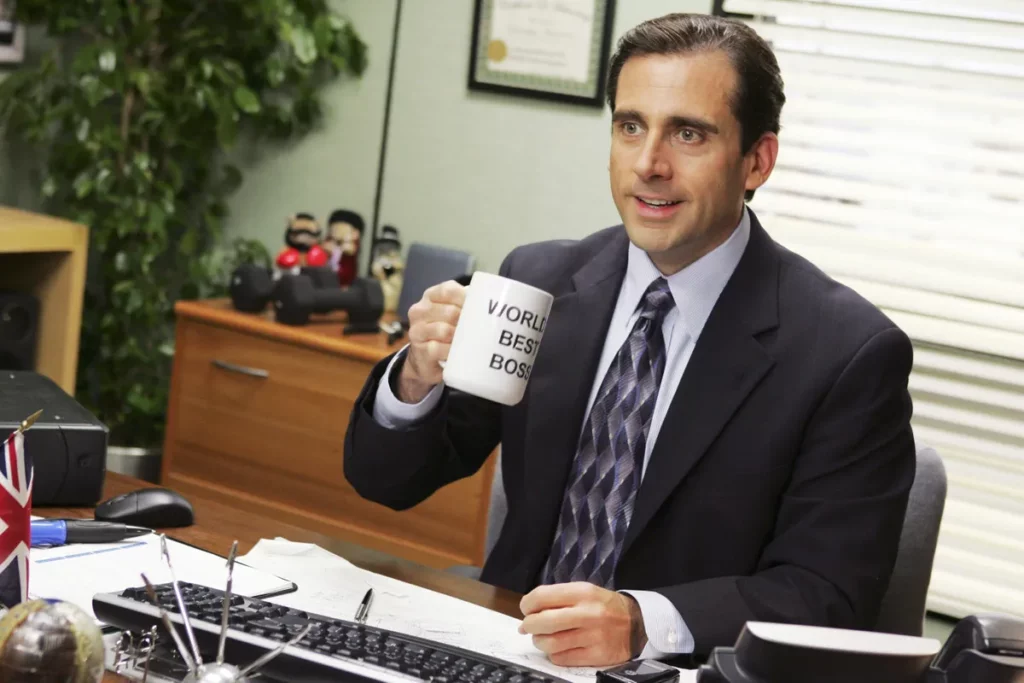 Cena da série The Office, em que o chefe Michael Scott está sentado de terno e segurando uma caneca de "melhor chefe do mundo".